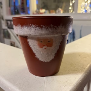May include: A terracotta flower pot painted with a white Santa Claus face. The pot has a white rim and a white beard on the Santa Claus.