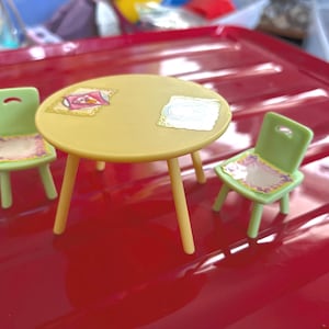 May include: A miniature yellow table with three legs and two green chairs. The chairs and table have decorative stickers on them. The set is on a red surface.
