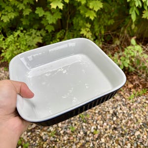 Puede incluir: Una fuente para hornear de cerámica blanca con un exterior negro acanalado. La fuente cuadrada se sostiene en una mano, con un fondo verde borroso de follaje. La fuente está vacía y parece sin usar.