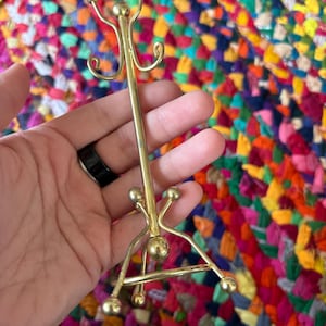 Può includere: Un piccolo attaccapanni in metallo color oro con un palo verticale e tre ganci curvi. L'attaccapanni ha una base a treppiede e dettagli a sfera decorativi. L'attaccapanni è alto circa 15 cm.