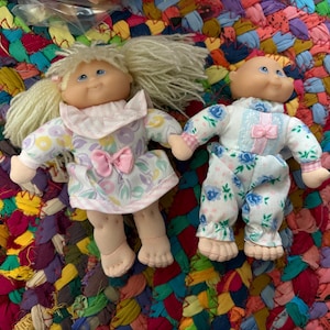 May include: Two vintage Cabbage Patch Kids dolls. One doll wears a floral dress with a pink bow, the other doll is in floral pajamas. Both dolls have blonde yarn hair and blue eyes. The dolls are on a colorful, textured surface.