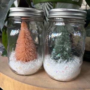 May include: Two glass mason jars with silver lids, each containing a miniature Christmas tree and white, sparkly snow. One tree is peach-colored, the other is green. The jars sit on a wooden surface.