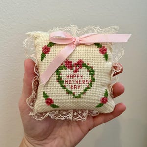 May include: A small white pillow with a pink ribbon tied around it. The pillow is embroidered with a green heart that says "Happy Mother's Day" and is surrounded by pink flowers.