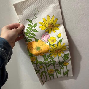May include: A white fabric tea towel with a floral pattern featuring yellow, pink, and green flowers. The flowers are stylized and have a retro look.
