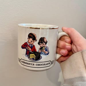 May include: White ceramic mug with a gold rim. The mug features a vintage illustration of two children eating a chocolate cake with the text "Hershey's Chocolate" on the bottom.