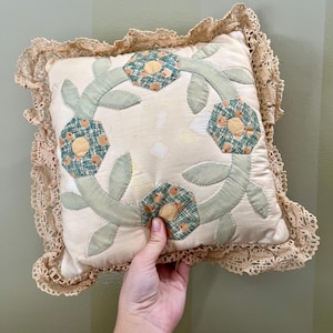 May include: A cream-colored pillow with a floral design. The pillow is quilted with green leaves and yellow and orange flowers. The pillow has a lace trim.