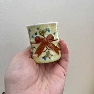 May include: A small, yellow ceramic cup with a brown bow and blue floral design.