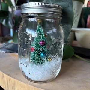 Könnte beinhalten: Ein klares Glas mit silbernem Deckel, das einen Miniatur-Weihnachtsbaum, bunte Ornamente und weißen Glitzer enthält. Das Glas steht auf einer Holzoberfläche, mit Grün im Hintergrund, was eine festliche Weihnachtsszene schafft.