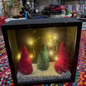 May include: A black framed shadow box with three miniature Christmas trees. Two red trees flank a green tree, all set on a bed of clear stones. String lights illuminate the scene, creating a festive display.