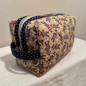 May include: A beige quilted cosmetic bag with a repeating pattern of sock monkey figures wearing red and white hats. The bag has a navy blue zipper and handle with white star accents. The bag is rectangular.