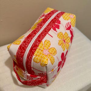 May include: A white zippered pouch with a red and yellow floral print. The pouch has a red handle and a zipper closure.