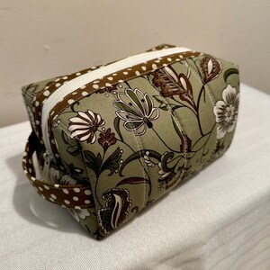 May include: A green quilted fabric cosmetic bag with a brown and white polka dot trim and a zipper closure. The bag has a floral pattern and a brown handle.