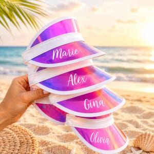 May include: A stack of four iridescent pink and purple sun visors with white trim. Each visor features a custom name printed in white script font: Marie, Alex, Gina, and Olivia. The visors have a shiny, reflective surface and a curved brim.