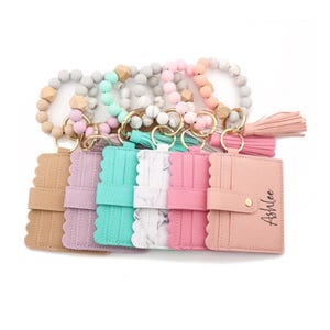 May include: Six card holders with scalloped edges and gold-tone snap closures, each attached to a beaded silicone bracelet with a tassel. The colors include tan, lavender, teal, white marble, pink, and peach. The peach holder features the name Ashlee.