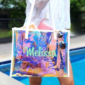 May include: A clear iridescent tote bag with white straps and the name "Melissa" printed in green. The bag is filled with items, including sunglasses.