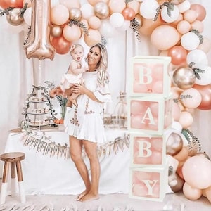 May include: A stack of clear boxes with the word "BABY" spelled out in white letters. Each box is filled with pink balloons. The boxes are in front of a wall decorated with a garland of peach, white, and gold balloons.