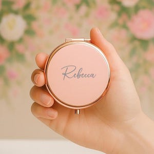 May include: A personalized, round compact mirror in a soft pink hue, held open to display the name "Rebecca" in elegant script. The compact has a rose gold trim. Other compacts in silver, gold, and black are visible.