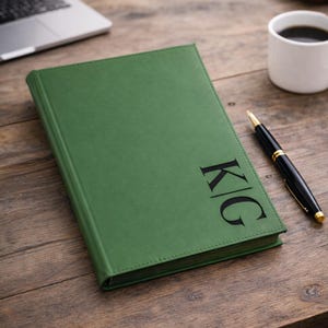 May include: A green leather journal with the initials "KG" in black lettering. A black and gold pen and a white mug of coffee sit nearby on a wooden desk. A laptop is visible in the background.