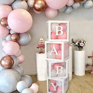 May include: A stack of four clear acrylic boxes with white frames, each containing pink and white balloons. The boxes are stacked to spell out the word "BABY". The boxes are decorated with a pink and white balloon garland.