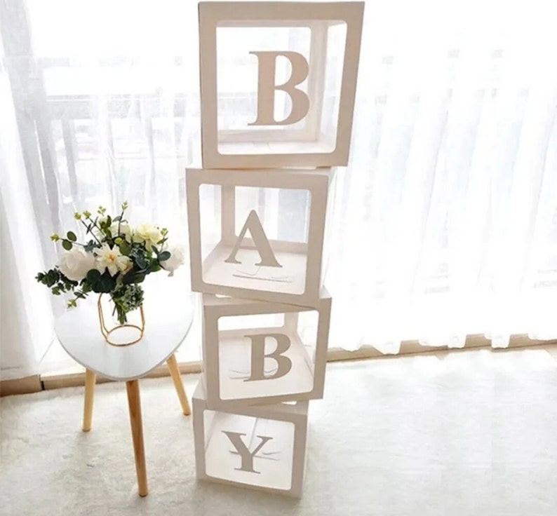 May include: Four white boxes stacked on top of each other, each with a clear front and a letter of the word "BABY" printed on the front. The boxes are made of cardboard and are decorated with a white ribbon.