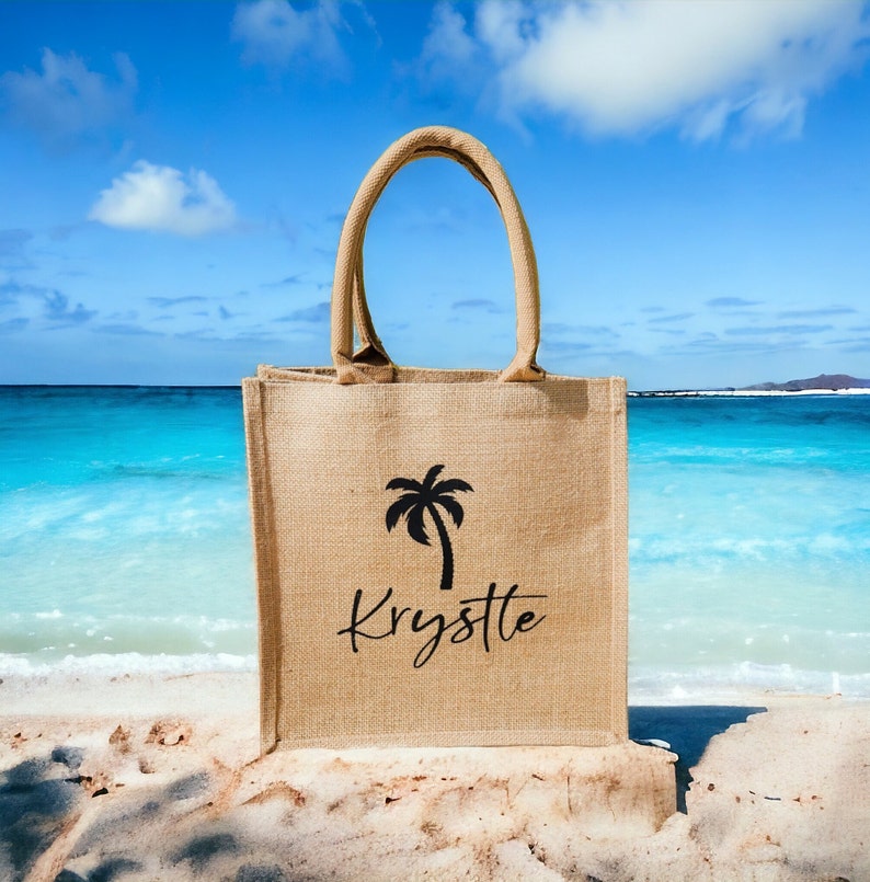 May include: A brown jute tote bag with a palm tree and the name "Krystle" printed on it. The bag is sitting on a sandy beach with blue water in the background.