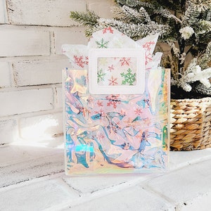 May include: A clear iridescent gift bag with a white frame and a window showing snowflake patterned tissue paper. The bag has a snowflake pattern.