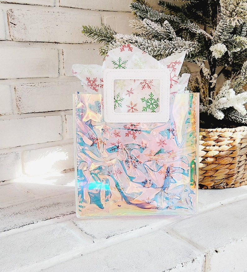 May include: A clear iridescent gift bag with a white frame and a window showing snowflake patterned tissue paper. The bag has a snowflake pattern.