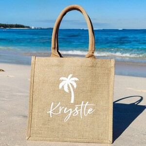 May include: A brown burlap tote bag with a white palm tree and the name "Krystle" printed on it. The bag is sitting on a sandy beach.
