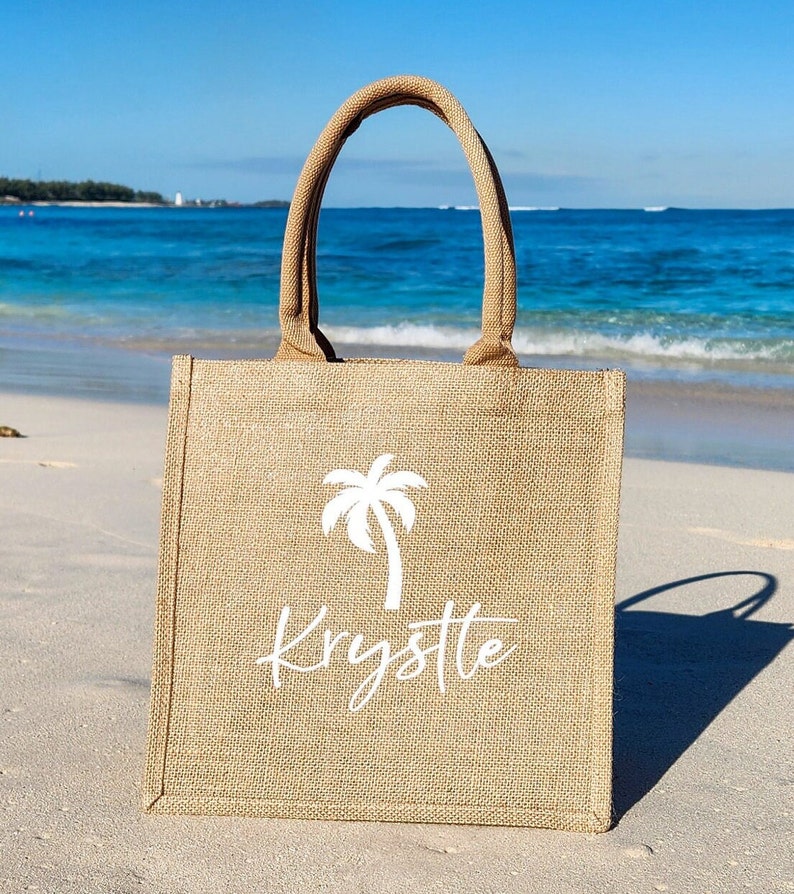 May include: A brown burlap tote bag with a white palm tree and the name "Krystle" printed on it. The bag is sitting on a sandy beach.