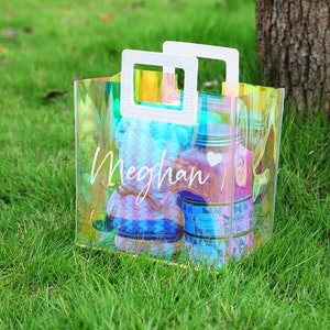 May include: A clear iridescent plastic tote bag with white handles and the name "Meghan" written on the front. The bag is filled with various items, including a blue and white patterned fabric.