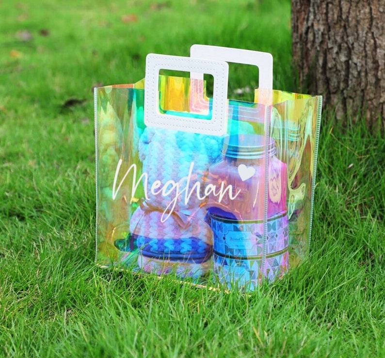 May include: A clear iridescent plastic tote bag with white handles and the name "Meghan" written on the front. The bag is filled with various items, including a blue and white patterned fabric.