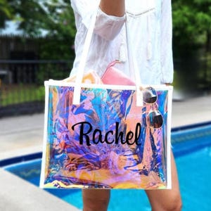 May include: A clear, iridescent tote bag with white handles and trim. The bag is personalized with the name "Rachel" in black script. Sunglasses hang from the handle, and a pink pouch is visible inside.