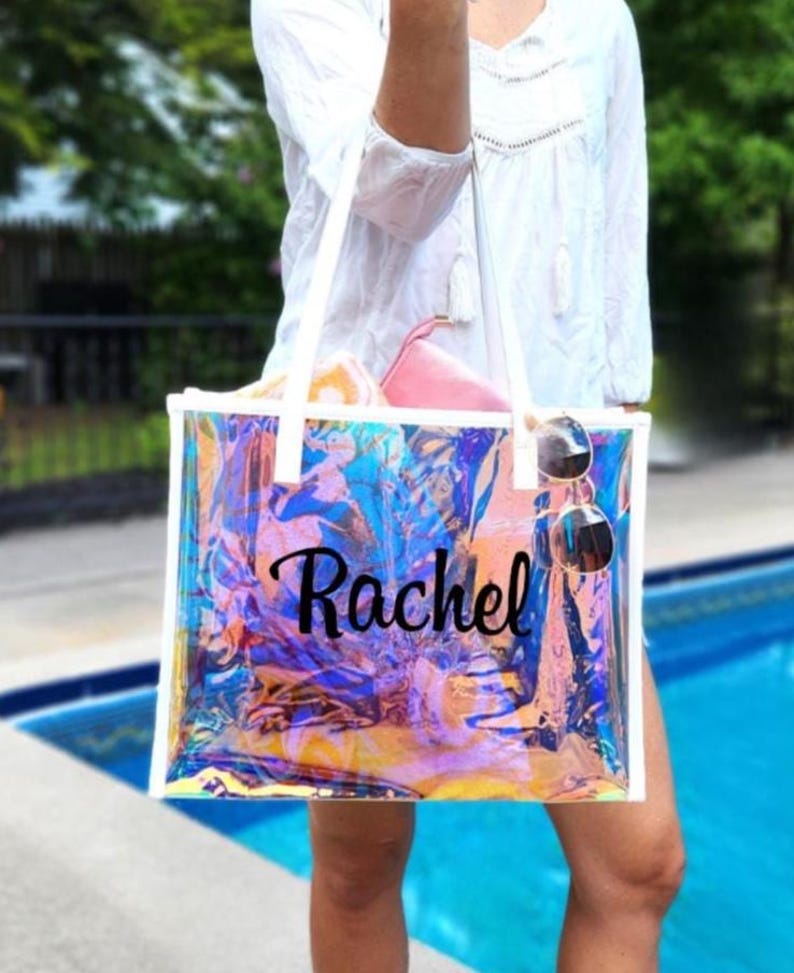 May include: A clear, iridescent tote bag with white handles and trim. The bag is personalized with the name "Rachel" in black script. Sunglasses hang from the handle, and a pink pouch is visible inside.
