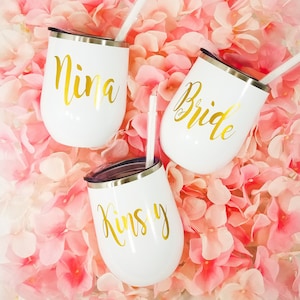 Puede incluir: Tres vasos de vino de acero inoxidable blanco con letras doradas. Los vasos est&aacute;n etiquetados con "Nina", "Bride" y "Kinsley".