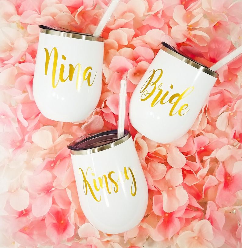 Puede incluir: Tres vasos de vino de acero inoxidable blanco con letras doradas. Los vasos est&aacute;n etiquetados con "Nina", "Bride" y "Kinsley".