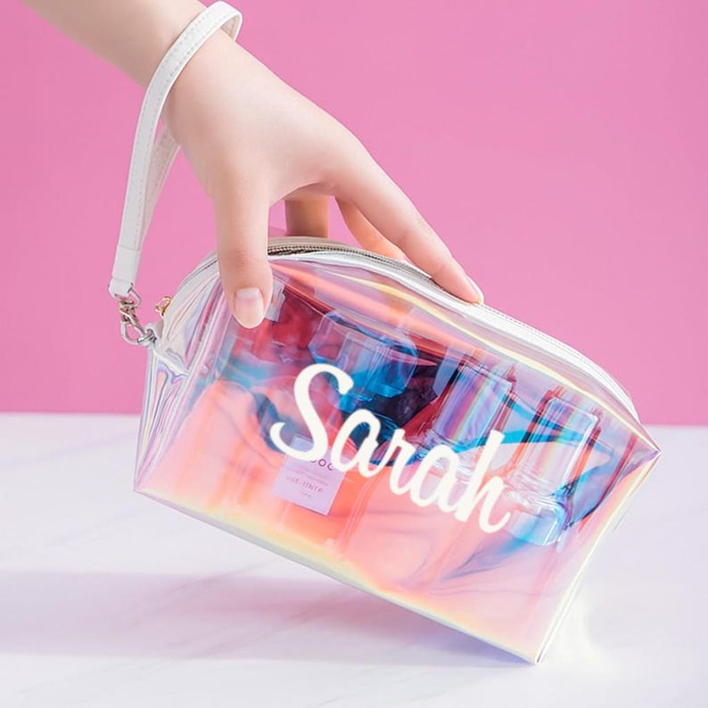 May include: Iridescent cosmetic bag with the name "Sarah" in white script. The clear bag has a white zipper and a white wrist strap. The bag reflects pink, blue, and yellow hues. The background is a solid pink.