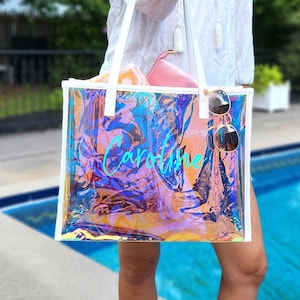May include: A clear, iridescent tote bag with white trim and handles. The bag has the name "Caroline" in turquoise script. Sunglasses hang from the side. A pink pouch and a towel are inside the bag. The background includes a pool.