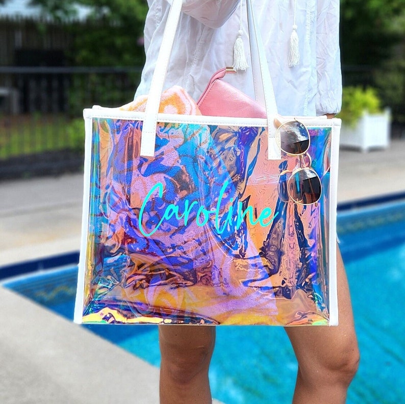 May include: A clear, iridescent tote bag with white trim and handles. The bag has the name "Caroline" in turquoise script. Sunglasses hang from the side. A pink pouch and a towel are inside the bag. The background includes a pool.