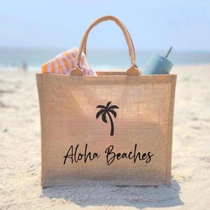 May include: A beige burlap tote bag with black palm tree and "Aloha Beaches" text. The bag is on a sandy beach.