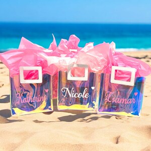May include: Three iridescent clear plastic tote bags with white handles and personalized names. The bags are on a sandy beach with pink tissue paper inside.