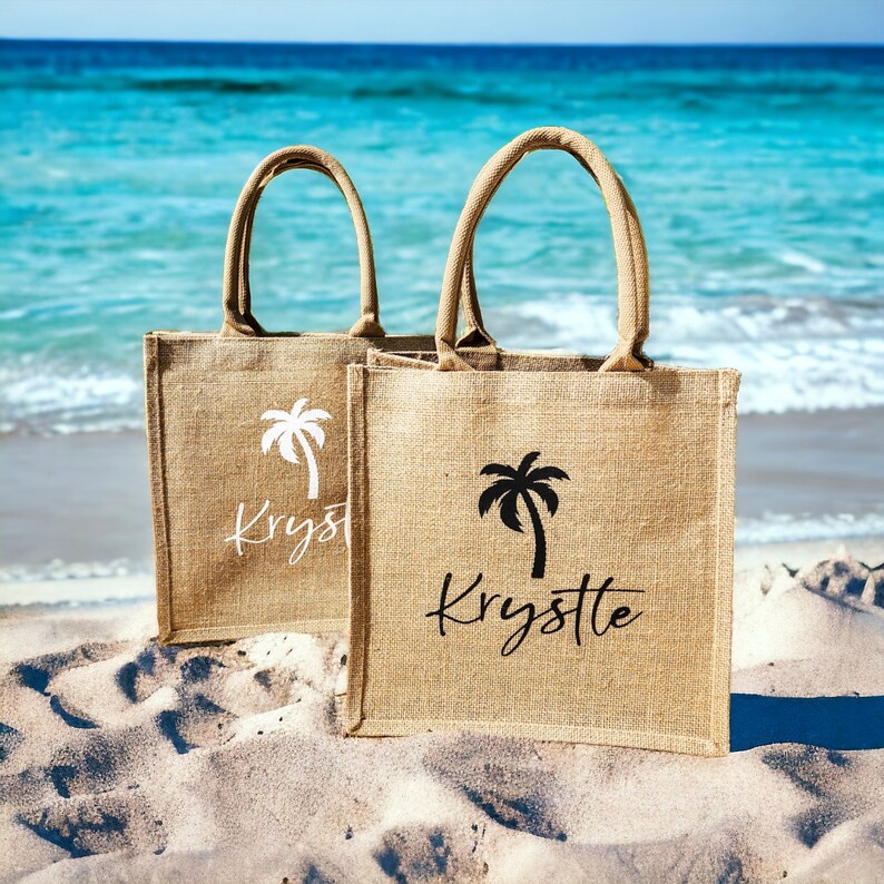 May include: Two brown burlap tote bags with palm tree designs and the name "Krystle" printed on them. The bags are sitting on a sandy beach.