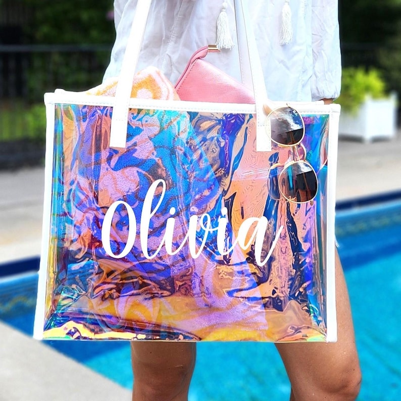 May include: A clear, iridescent tote bag with white trim and handles. The bag features the name "Olivia" in white script. Sunglasses hang from the handle. A pink pouch and a towel are visible inside the bag. The bag is near a swimming pool.