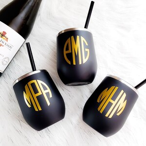 Puede incluir: Tres vasos de vino de acero inoxidable negro con iniciales de monograma doradas. Los vasos tienen un borde plateado y est&aacute;n sobre una superficie blanca.