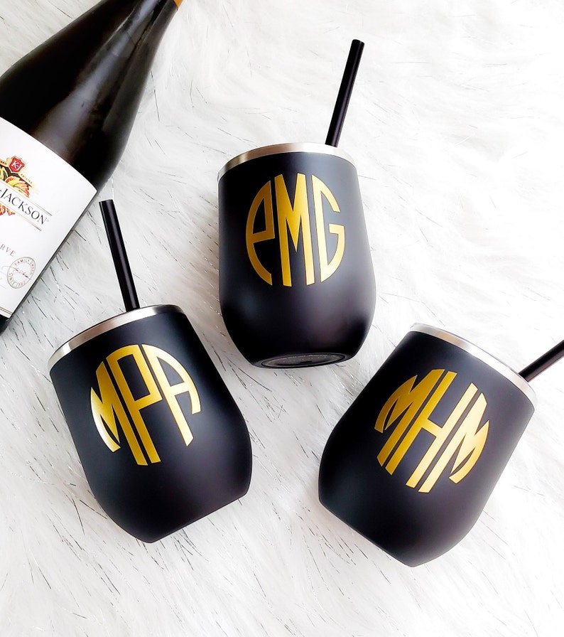 Puede incluir: Tres vasos de vino de acero inoxidable negro con iniciales de monograma doradas. Los vasos tienen un borde plateado y est&aacute;n sobre una superficie blanca.