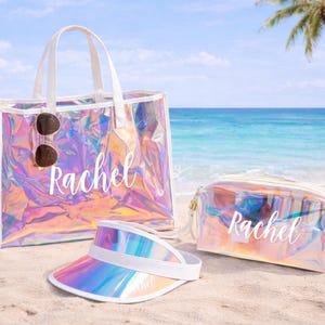May include: A clear iridescent tote bag, cosmetic bag, and visor sit on a sandy beach. The tote bag has white handles and sunglasses attached. The name "Rachel" is written on each item in white script. The visor has a white band.