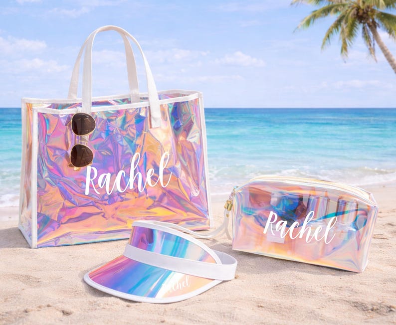 May include: A clear iridescent tote bag, cosmetic bag, and visor sit on a sandy beach. The tote bag has white handles and sunglasses attached. The name "Rachel" is written on each item in white script. The visor has a white band.
