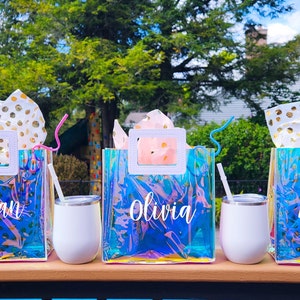 May include: Three iridescent clear plastic bags with white handles and gold polka dot tissue paper inside. Each bag has a name written on it: Meghan, Olivia, and Angela. Each bag is paired with a white tumbler and a straw.