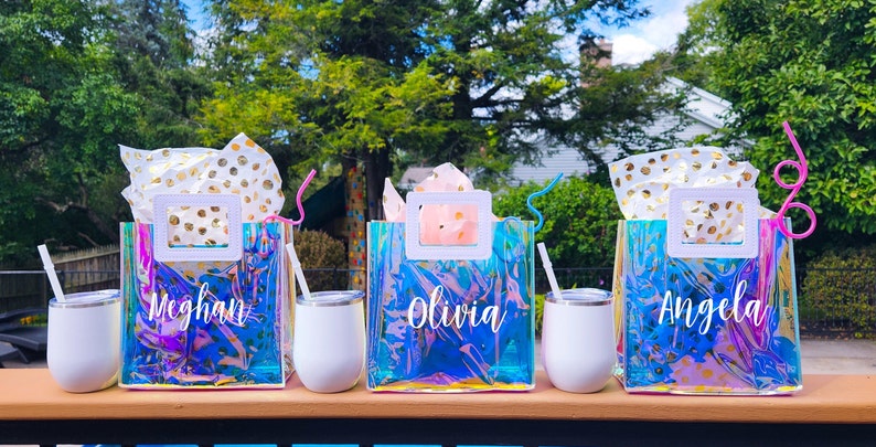 May include: Three iridescent clear plastic bags with white handles and gold polka dot tissue paper inside. Each bag has a name written on it: Meghan, Olivia, and Angela. Each bag is paired with a white tumbler and a straw.