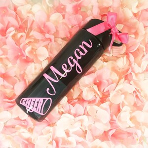 K&ouml;nnte beinhalten: Schwarze Wasserflasche mit einem rosa Band und dem Namen "Megan" in rosa Schrift. Die Flasche hat ein rosa Cheerleader-Megaphon-Motiv.