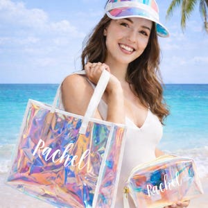 May include: A holographic tote bag and matching cosmetic bag with the name "Rachel" in white script. The bags are iridescent and appear to change colors. A rainbow visor is worn on the head. The background is a beach scene with blue water and sky.